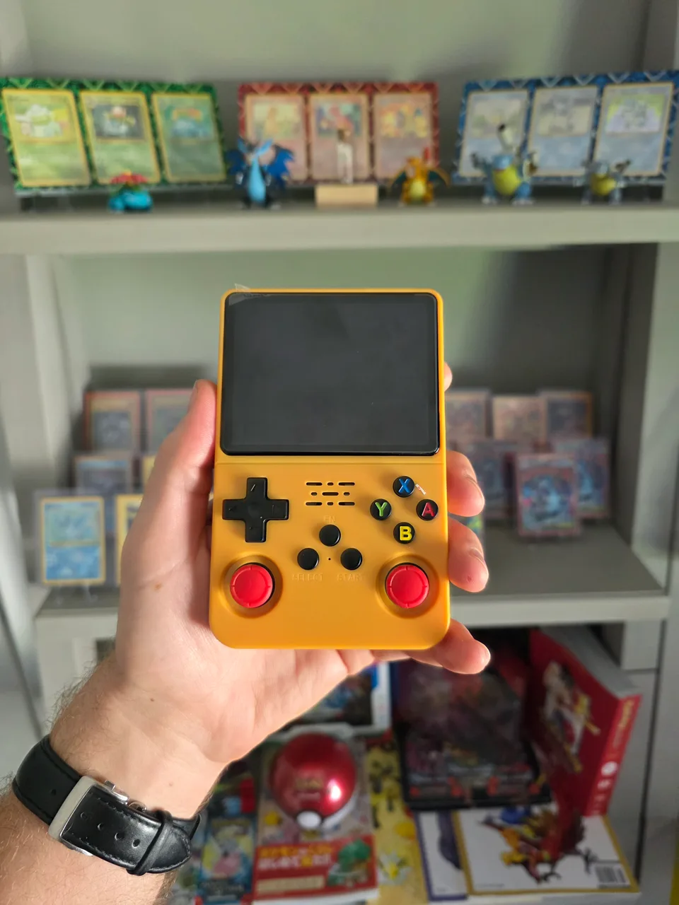 A yellow R36S handheld console, screen off, held in one hand in front of a shelf of Pokemon cards and figures.